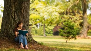 woman sitting outside under a tree reading a book 10 Way to reduce stress naturally